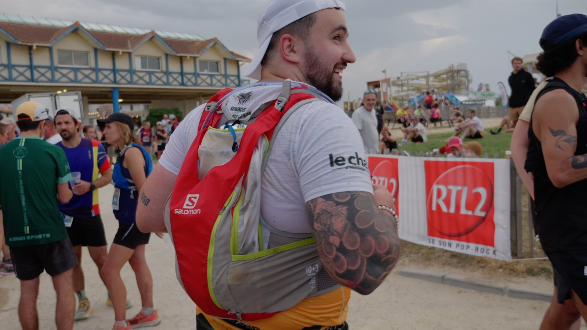 Coureur tatoué de dos avec sac Salomon devant les banderoles RTL2